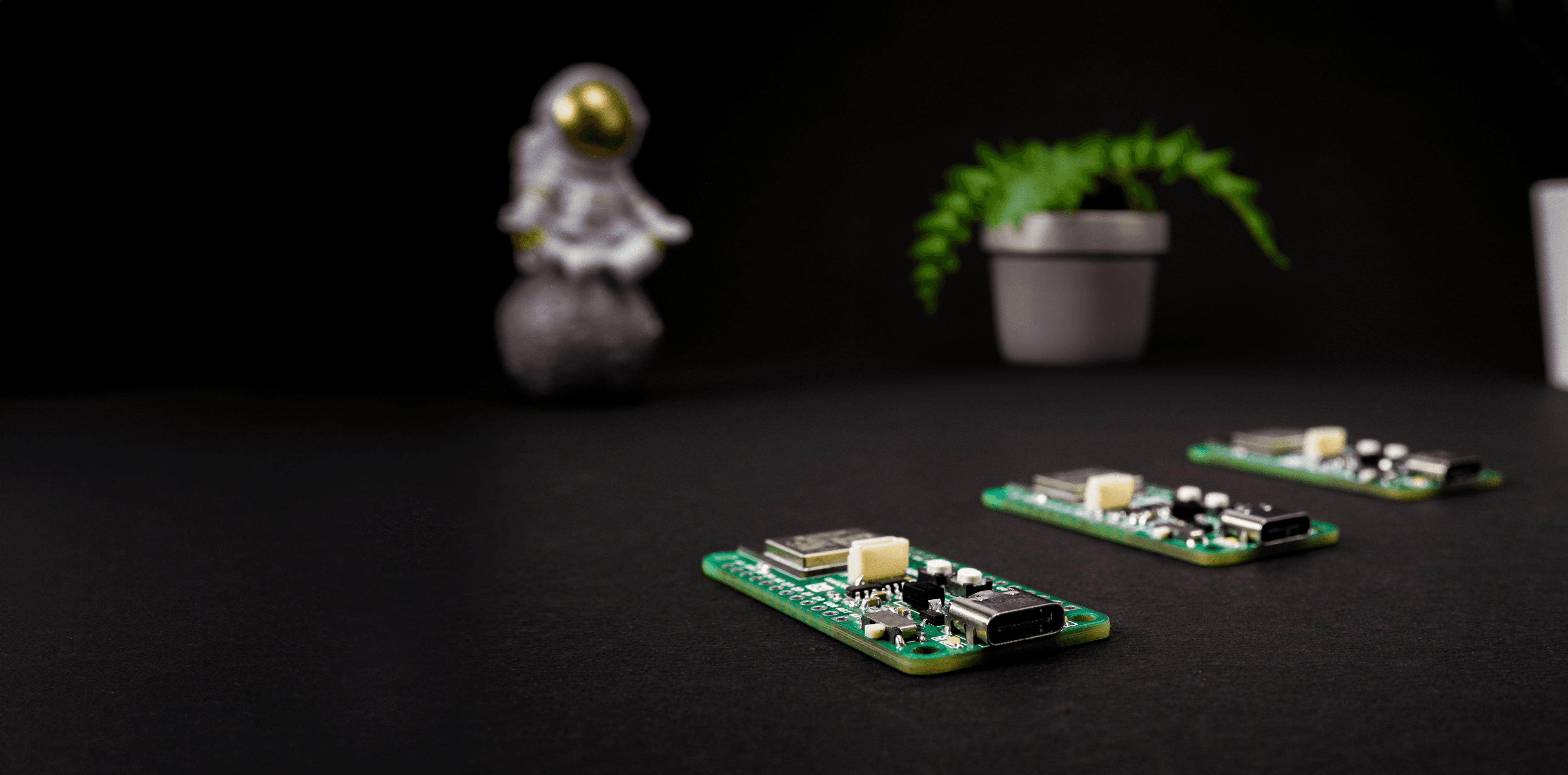 Two green microcontroller PCBs on a dark desk with miniature astronaut figurine and potted plant.