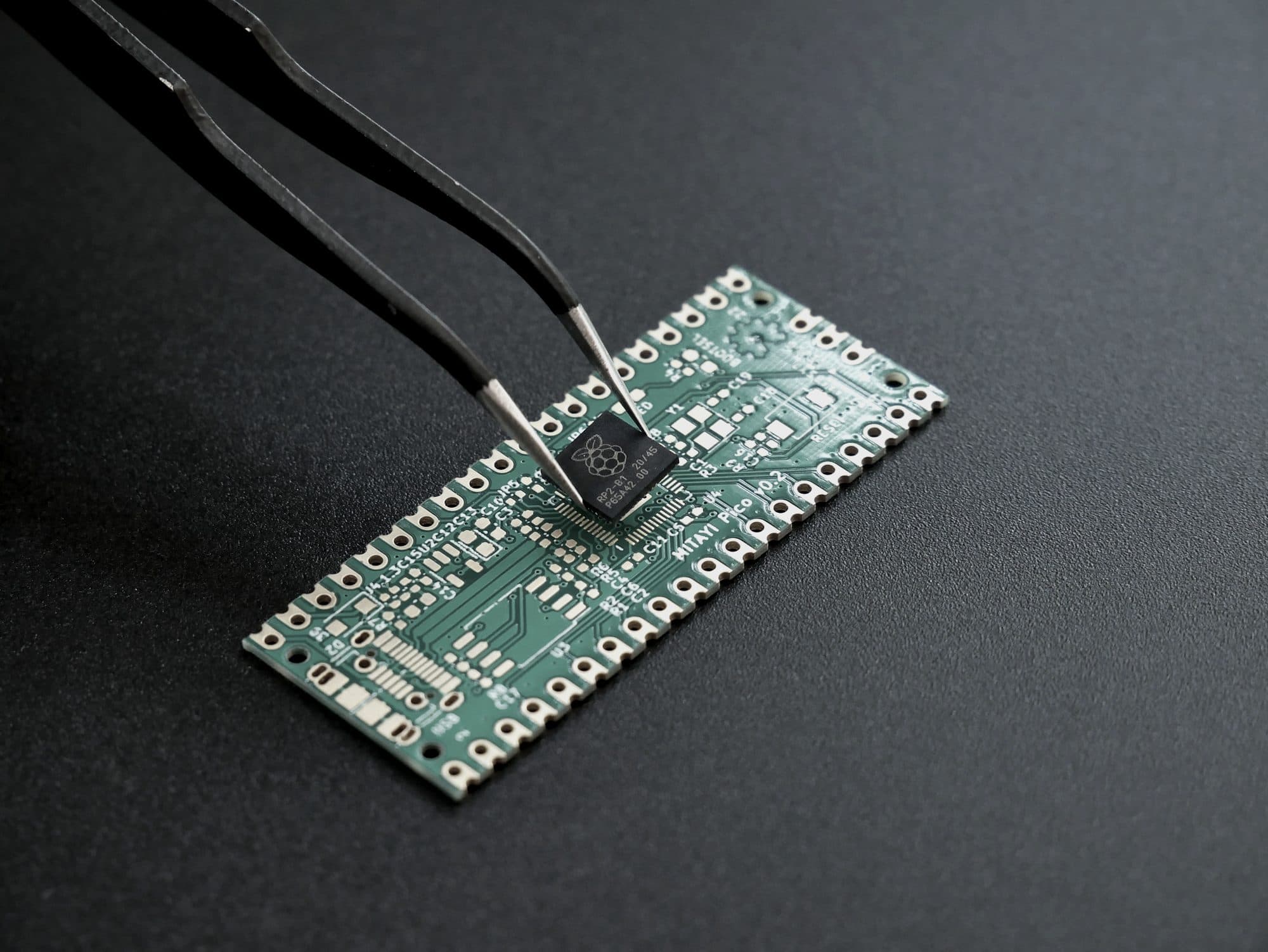 Tweezers lowering a microcontroller chip onto a bare prototype circuit board marked v0.2.